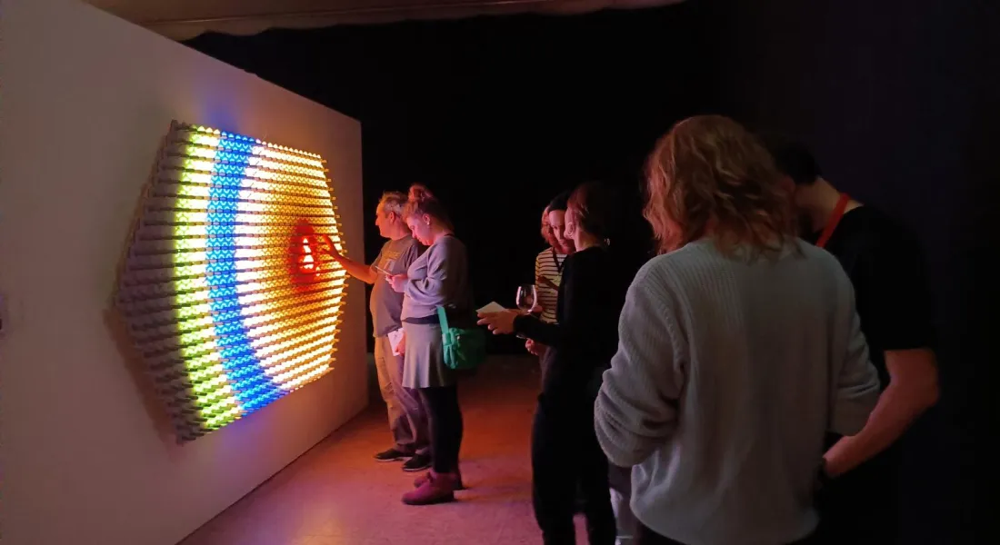 Mehrer Ausstellungsbesucher*innen stehen um ein Kunstwerk aus LEDs an der Wand; einige davon berühren das Werk