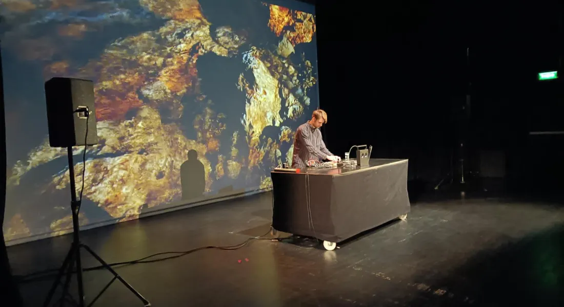 Ein Medienkünstler, an einem Tisch stehend, über seine Konsolen gebeugt, im Hintergrund eine Videprojektion von Steinen im Wasser